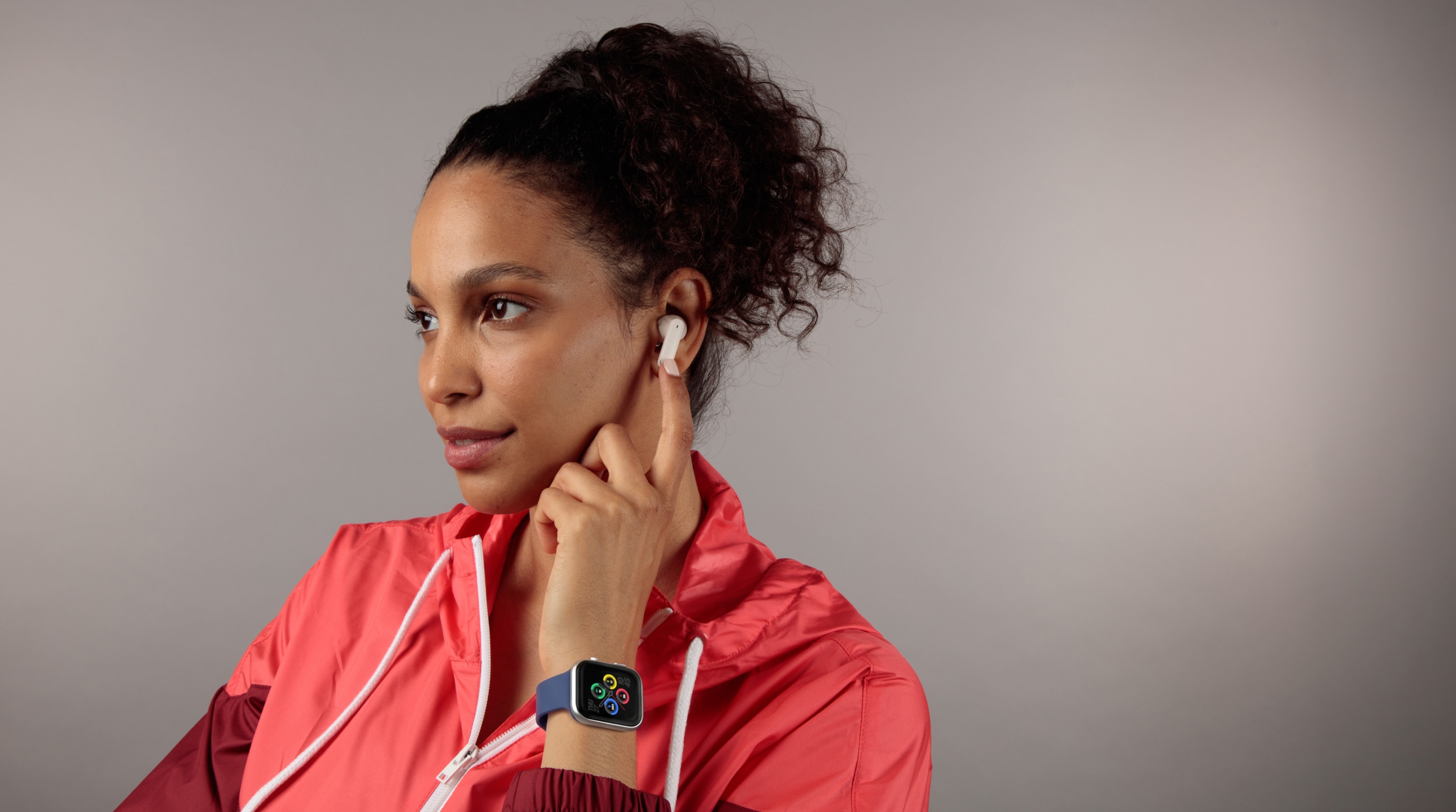 Woman wearing a red jacket and smartwatch, holding an earbud to her ear against a gray background